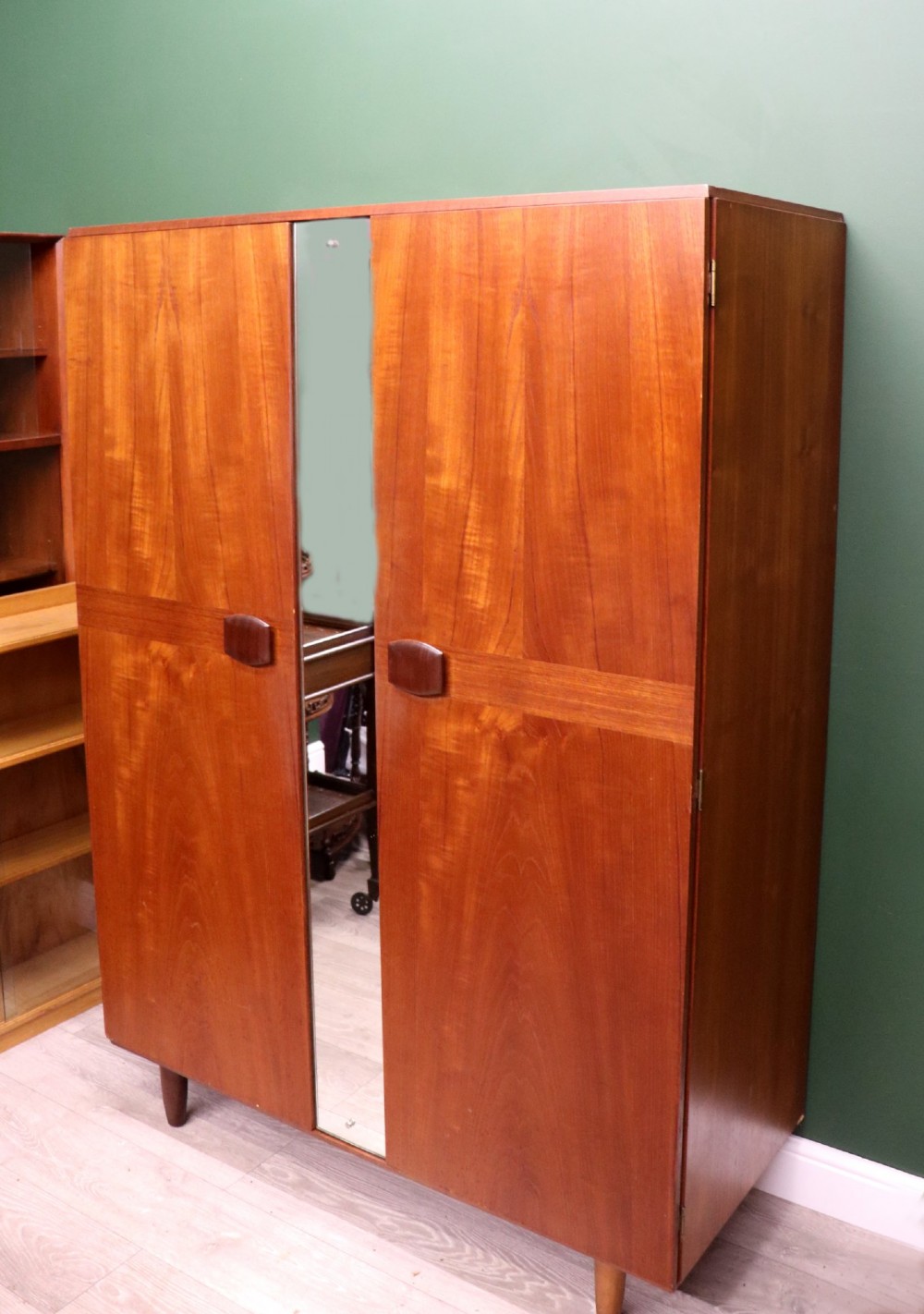 A mid-century teak wood wardrobe featuring two doors with rectangular wooden handles, short tapered legs, and a narrow central full-length mirror. View II