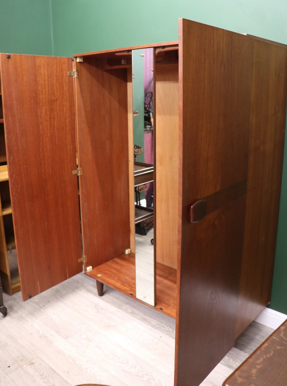 A mid-century teak wood wardrobe featuring two doors with rectangular wooden handles, short tapered legs, and a narrow central full-length mirror. View III