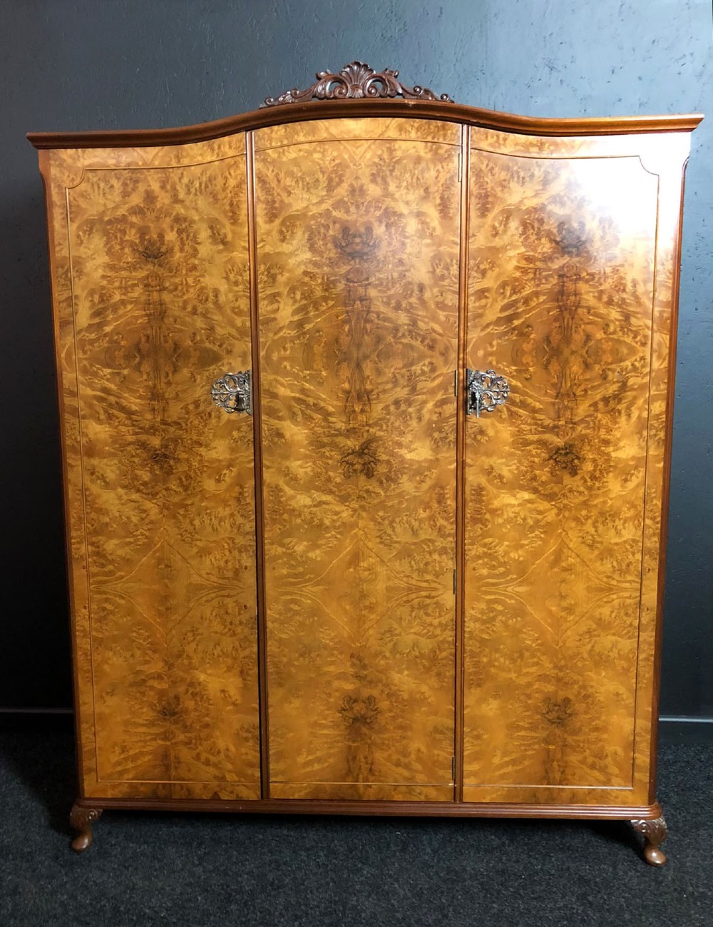 A large Queen Anne revival three-door wardrobe crafted with highly figured burr walnut veneer, featuring carved cresting and decorative metal escutcheons.