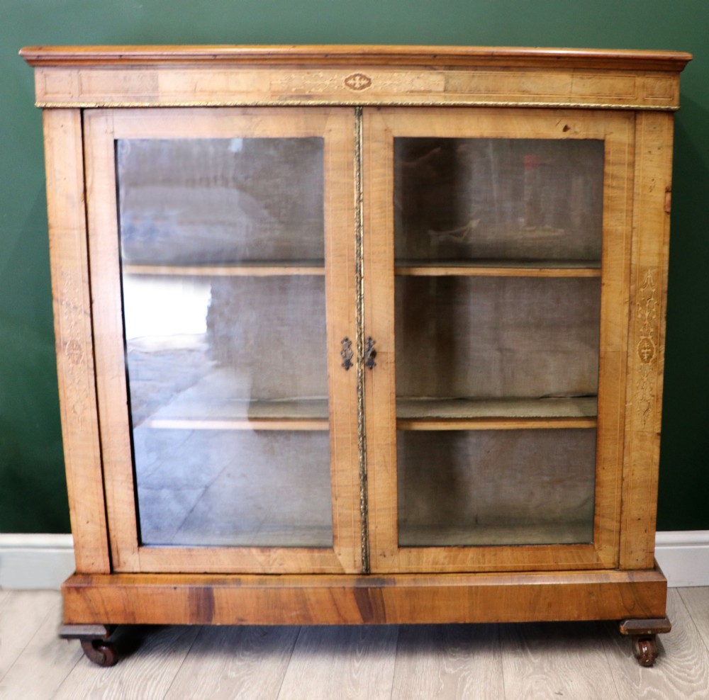 A mid-Victorian low walnut veneered bookcase featuring two glass glazed doors, marquetry inlay along the frieze, and brass trim detailing.