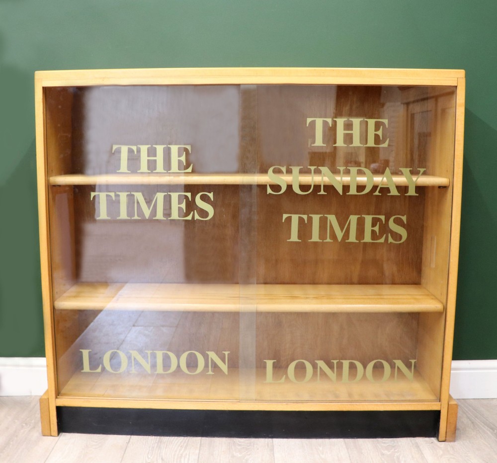 A low mid-century light oak bookcase with sliding glass doors featuring printed gold typography reading The Times and The Sunday Times London.