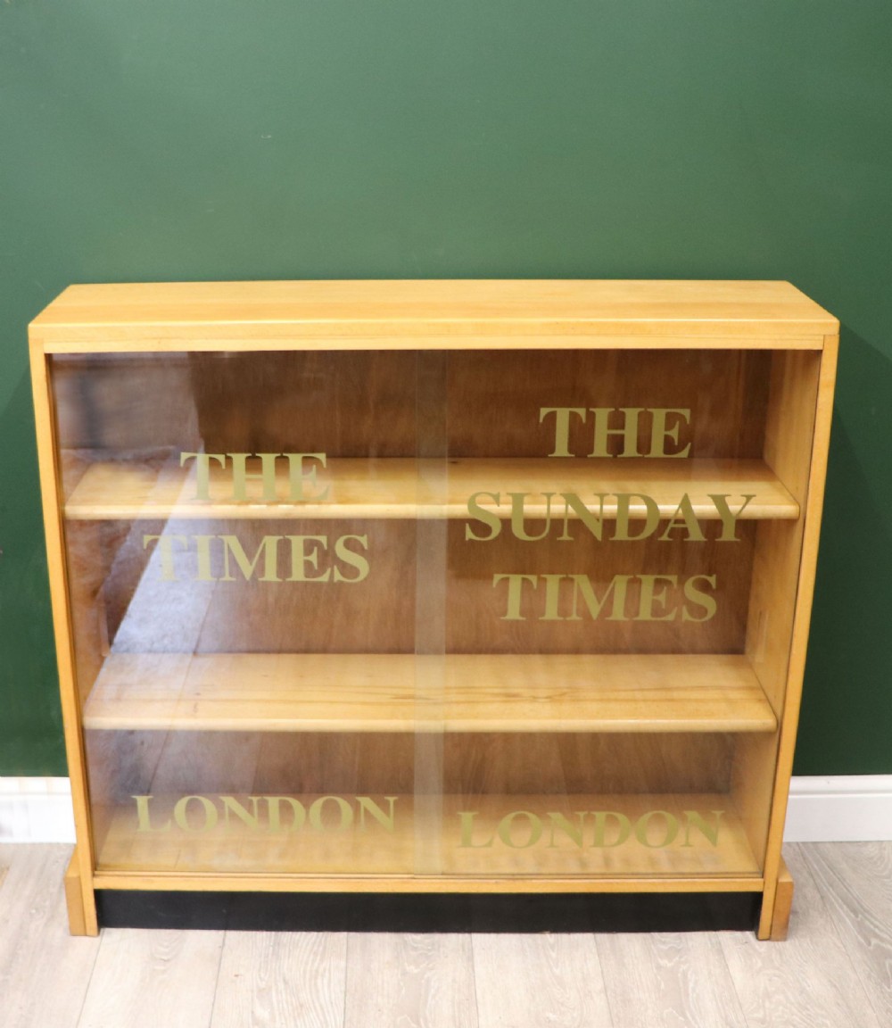 A low mid-century light oak bookcase with sliding glass doors featuring printed gold typography reading The Times and The Sunday Times London. View II