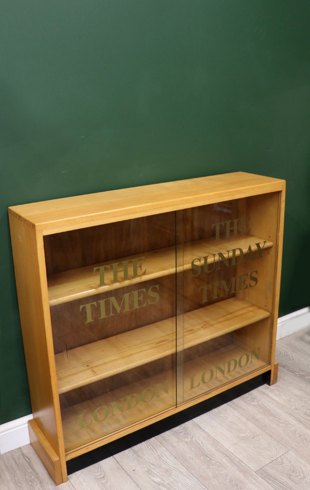 A low mid-century light oak bookcase with sliding glass doors featuring printed gold typography reading The Times and The Sunday Times London. View V