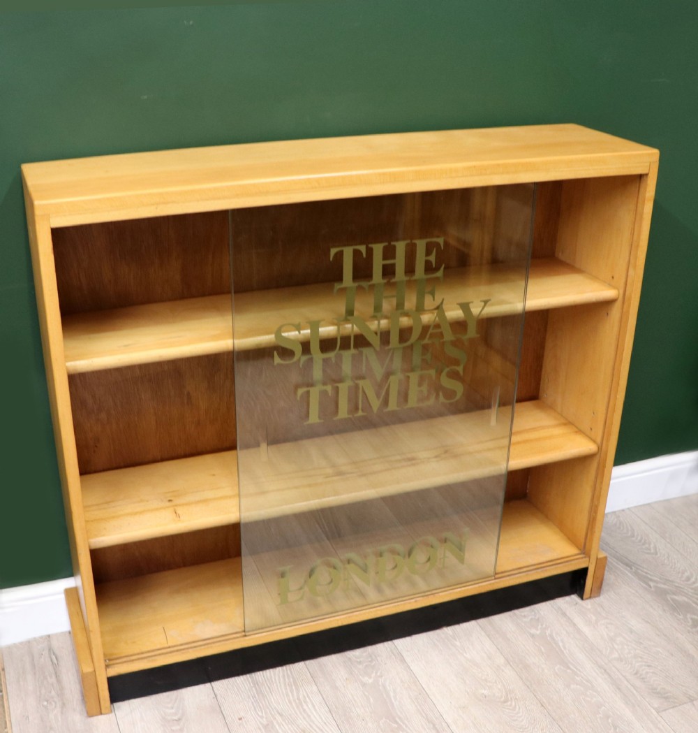 A low mid-century light oak bookcase with sliding glass doors featuring printed gold typography reading The Times and The Sunday Times London. View VI