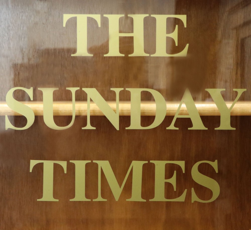 A low mid-century light oak bookcase with sliding glass doors featuring printed gold typography reading The Times and The Sunday Times London. View X