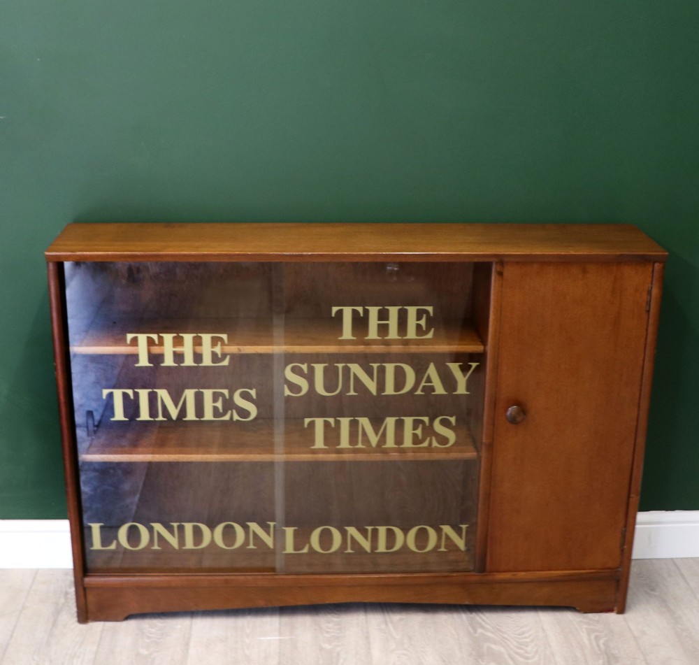 A low mid-century teak bookcase with two sliding glass doors featuring printed lettering alongside a single solid teak cupboard door.
