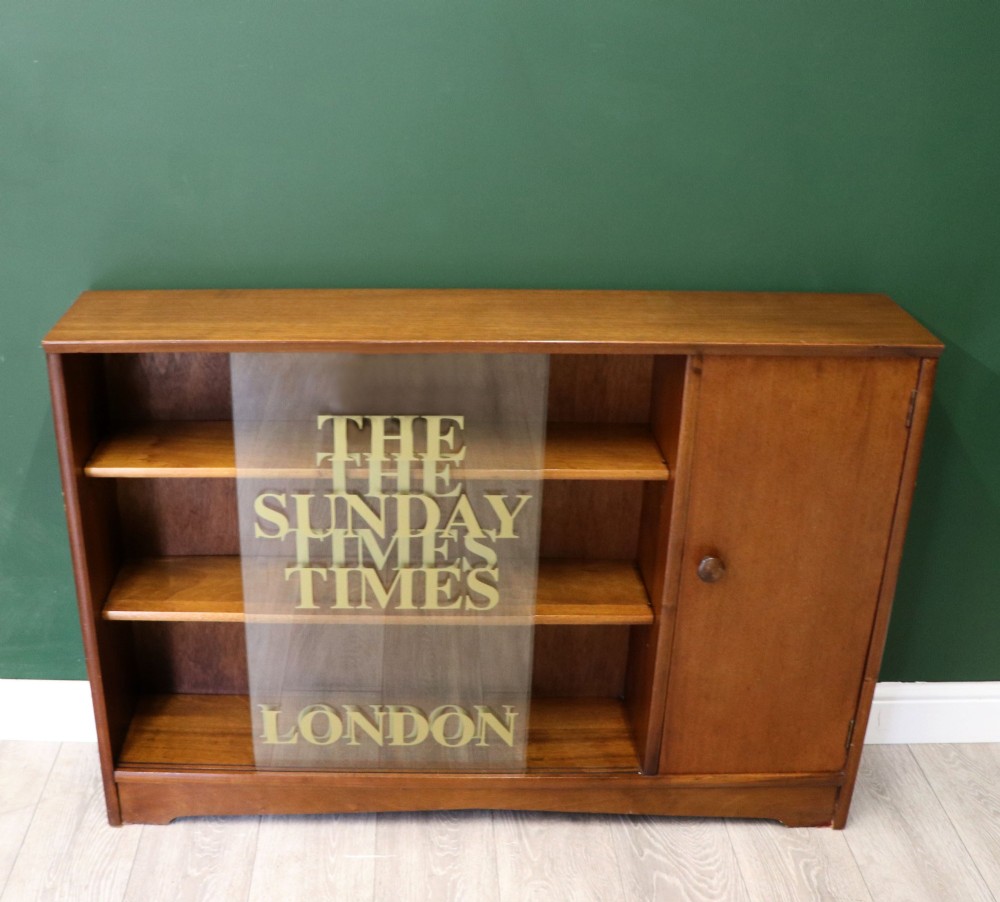 A low mid-century teak bookcase with two sliding glass doors featuring printed lettering alongside a single solid teak cupboard door. View II