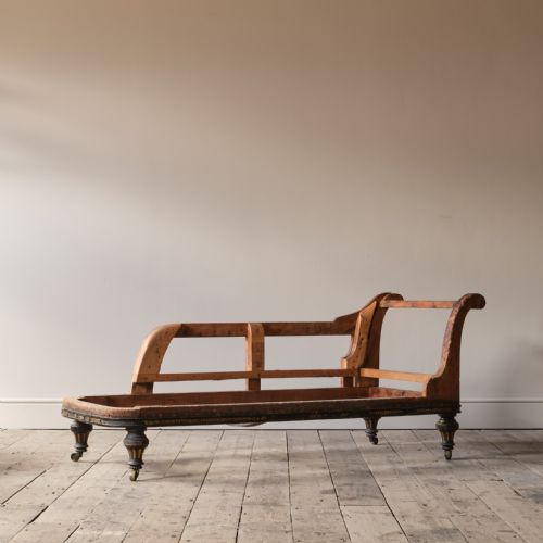 fig 1. An un-upholstered Victorian wooden chaise longue frame featuring a curved backrest, ebonised seat rail details, and turned legs on brass castors.