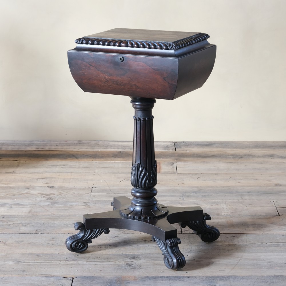 A dark rosewood Regency teapoy featuring gadrooned moulding, a carved column pedestal, and four scrolled legs.