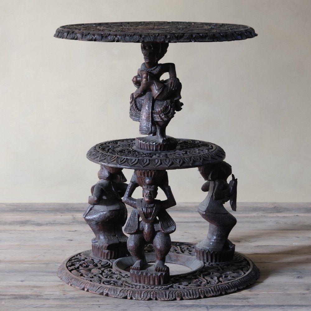 A late 19th-century Burmese dark teak two-tier side table with hand-carved mythological figure supports and circular foliate-carved surfaces.