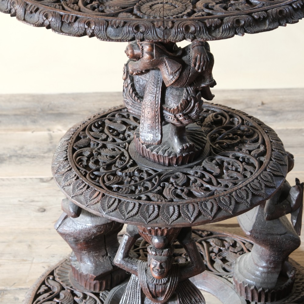 A late 19th-century Burmese dark teak two-tier side table with hand-carved mythological figure supports and circular foliate-carved surfaces. View II