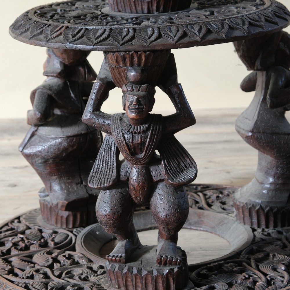 A late 19th-century Burmese dark teak two-tier side table with hand-carved mythological figure supports and circular foliate-carved surfaces. View V
