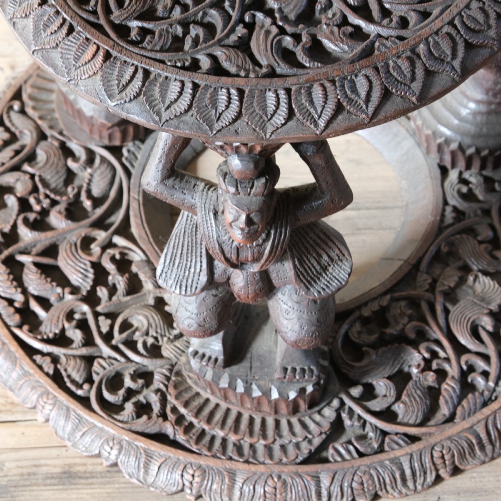 A late 19th-century Burmese dark teak two-tier side table with hand-carved mythological figure supports and circular foliate-carved surfaces. View VI