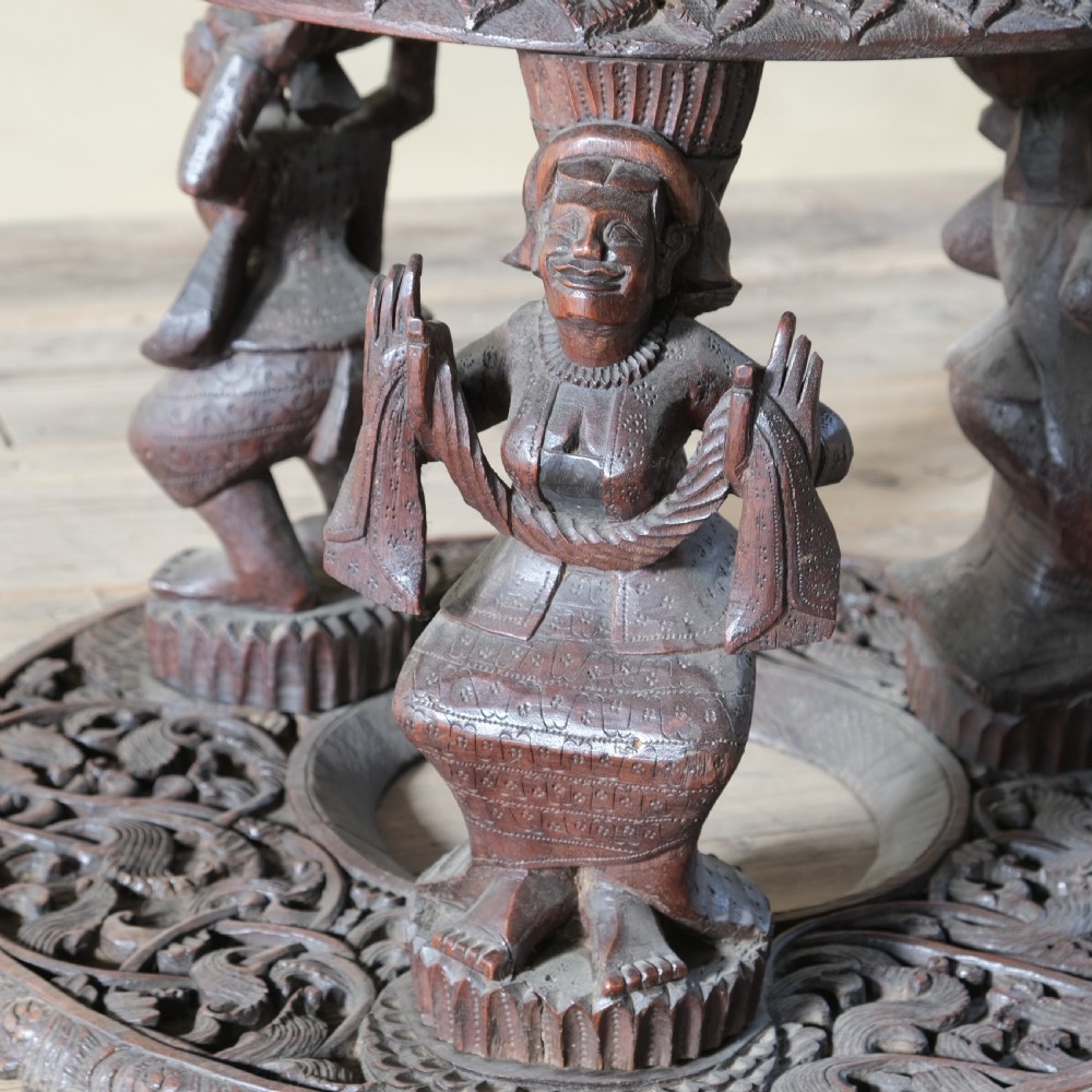 A late 19th-century Burmese dark teak two-tier side table with hand-carved mythological figure supports and circular foliate-carved surfaces. View X