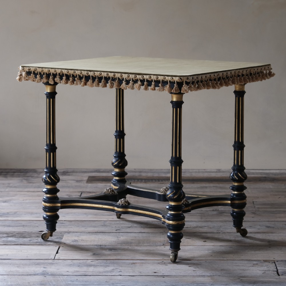 An Aesthetic Movement ebonised and parcel-gilt square table featuring fluted turned legs, a tassel fringe skirt, and brass castors.