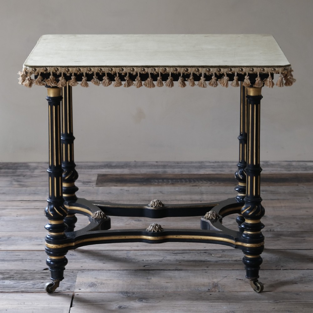 An Aesthetic Movement ebonised and parcel-gilt square table featuring fluted turned legs, a tassel fringe skirt, and brass castors. View II