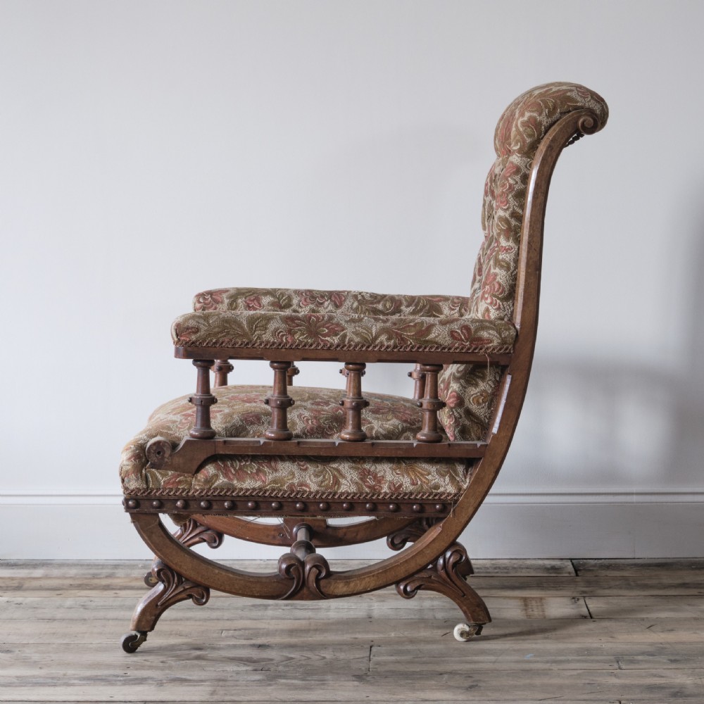 A profile view of a 19th-century Gothic Revival open armchair with a carved walnut frame, turned spindles under padded armrests, and floral tapestry upholstery.