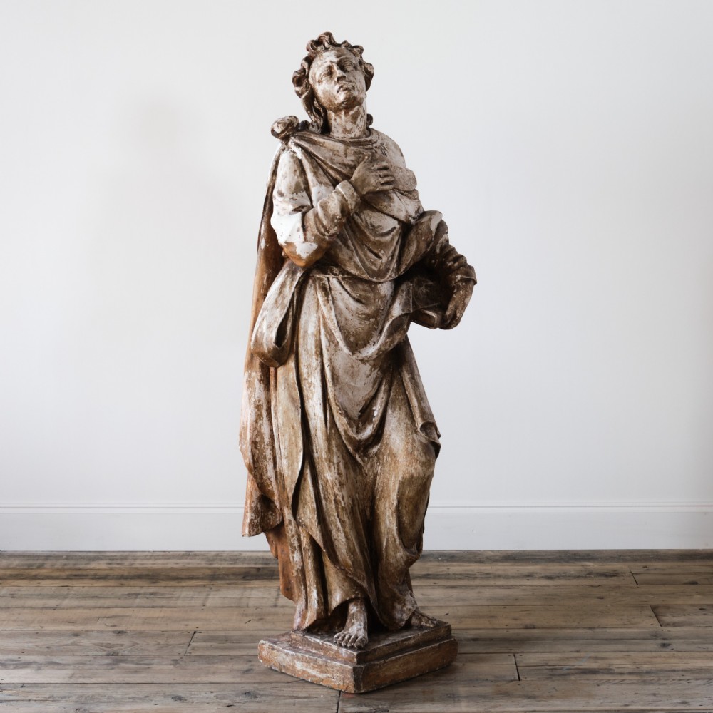 A large freestanding ecclesiastical plaster statue of a saint wearing draped robes, resting on a square pedestal with a distressed brown patinated surface.