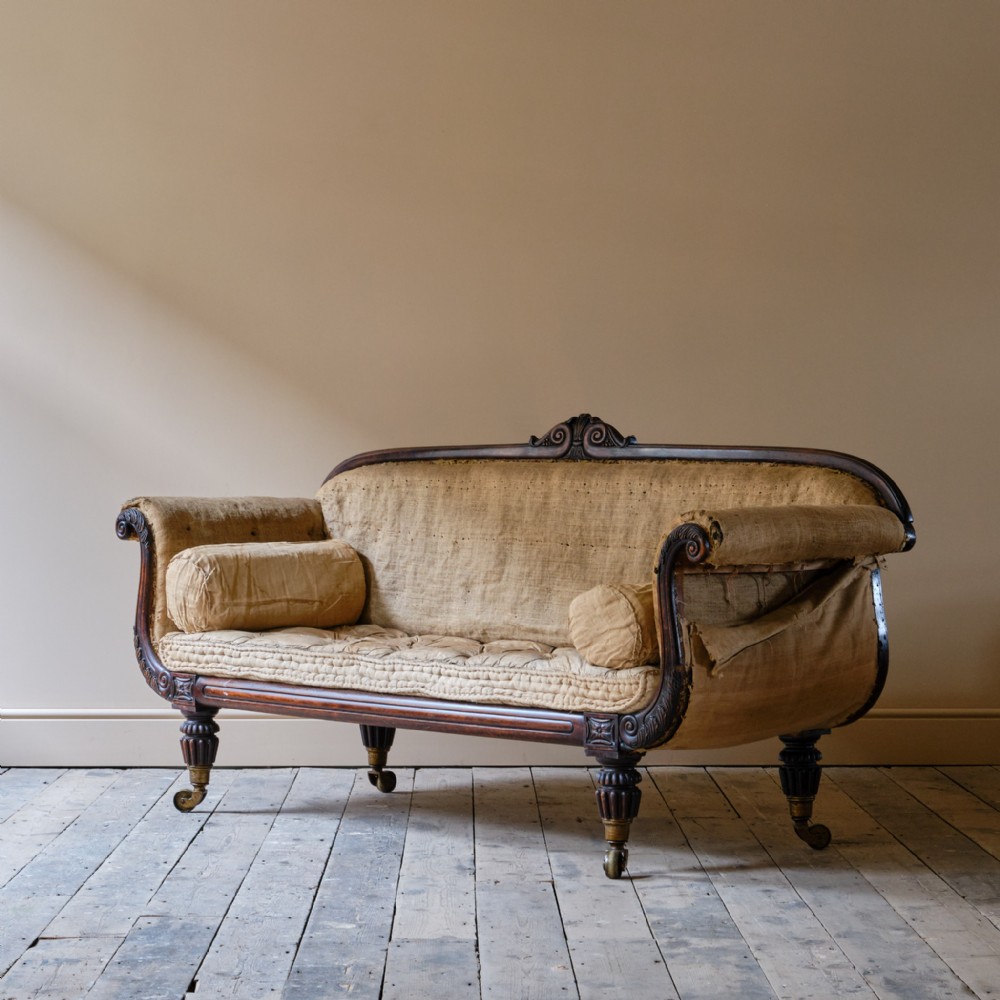 An early nineteenth-century Regency scroll-arm sofa featuring a simulated rosewood finish, carved cresting, reeded legs on brass castors, and neutral upholstery.
