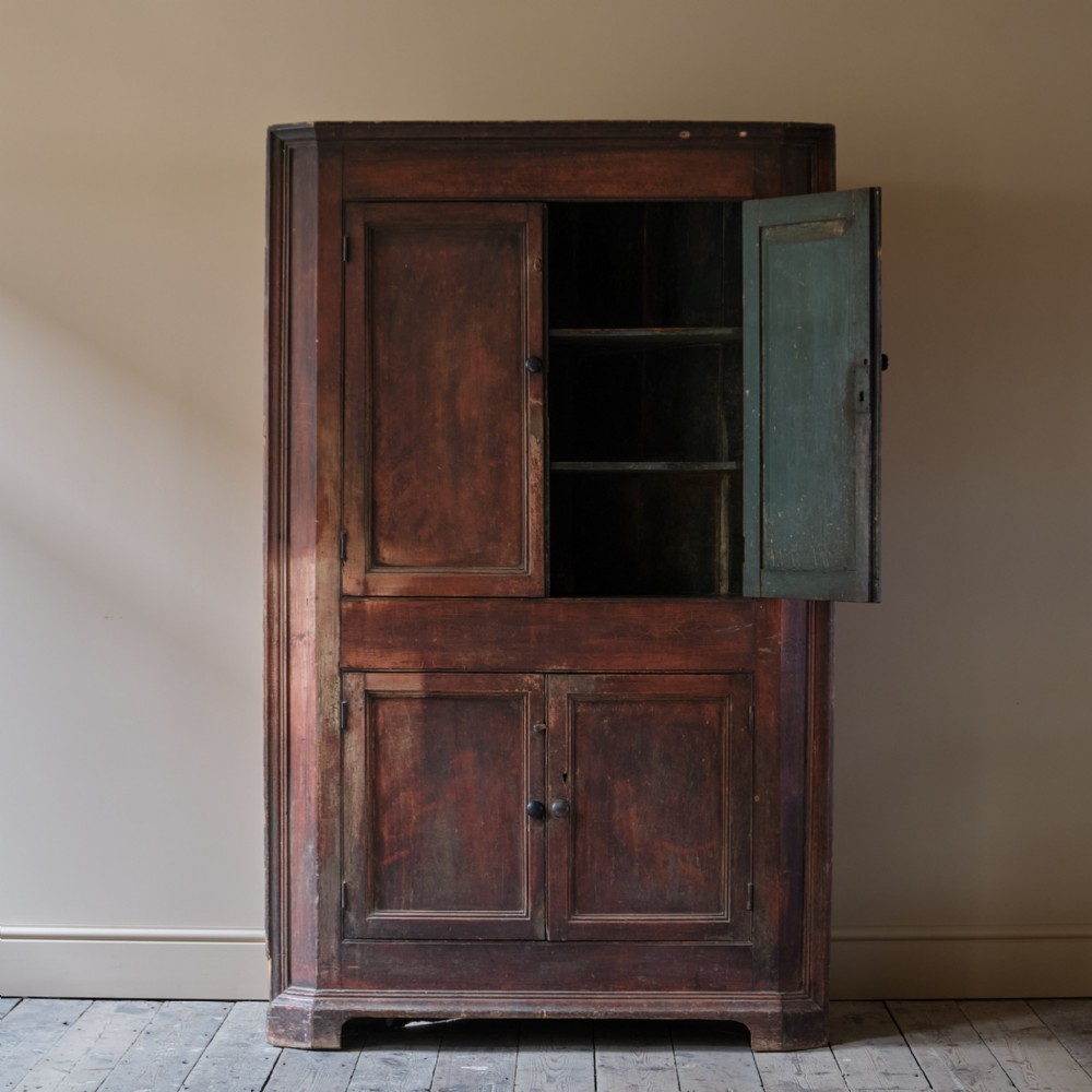 A tall freestanding Georgian corner cupboard featuring a worn dark red painted exterior, four panelled doors, and an open upper section revealing a green painted interior.