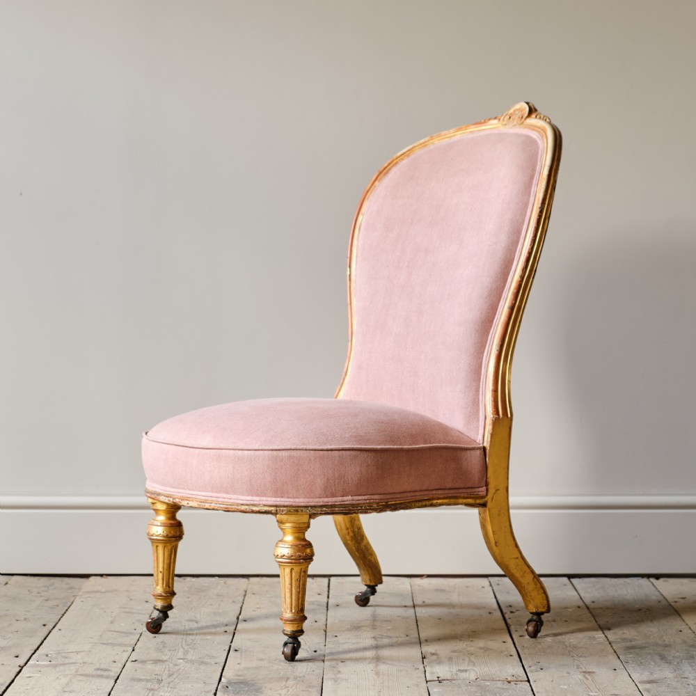 A 19th-century armless slipper chair featuring a carved giltwood frame with turned front legs, brass castors, and dusty pink velvet upholstery.