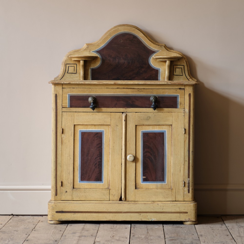 A 19th-century pine cupboard painted in ochre yellow featuring faux-marbled reddish-brown door and drawer panels, framed with blue paint lines and topped with an arched gallery shelf.