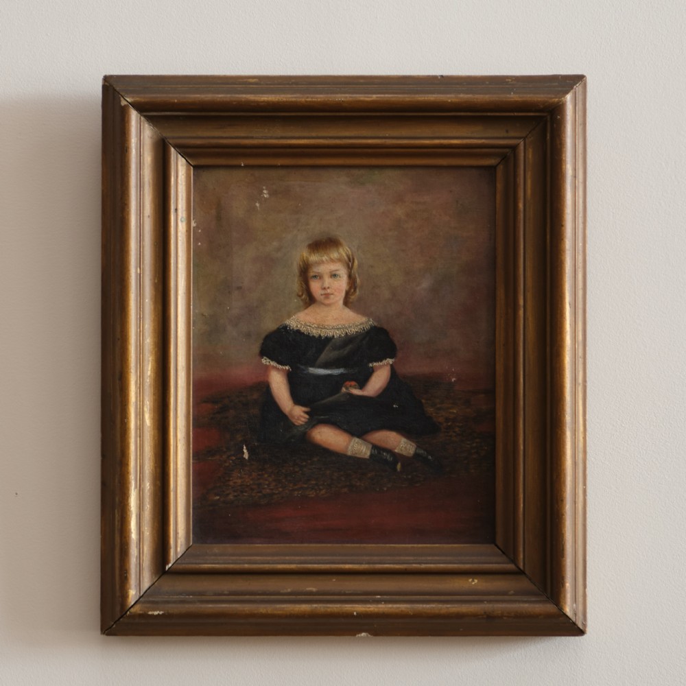A nineteenth-century naive oil painting on canvas depicting a seated child in a dark lace-trimmed dress, framed in a moulded gilt timber frame.