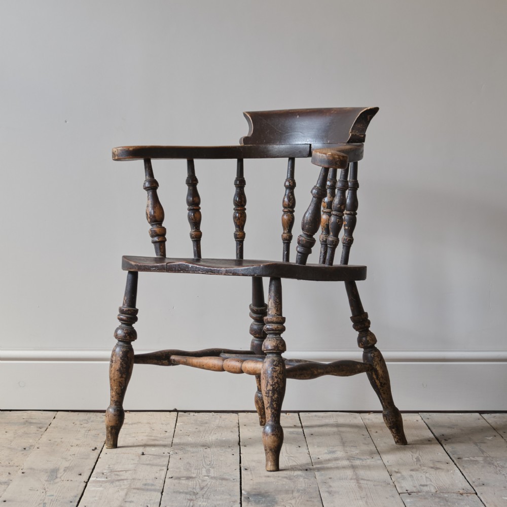A nineteenth-century dark brown elm smokers bow chair with turned spindles, a solid saddle seat, and turned legs joined by a stretcher base.