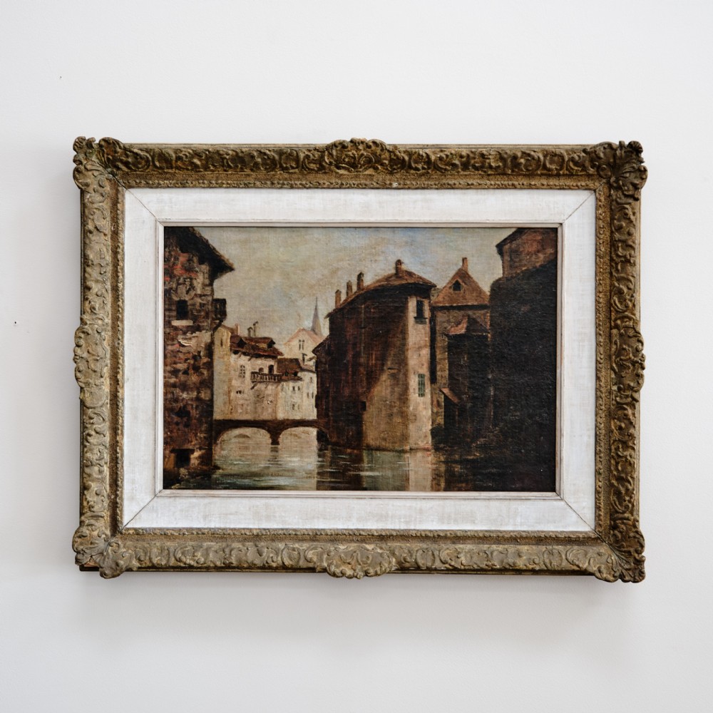 A 19th-century French oil painting on canvas depicting a quiet canal town scene with stone buildings, set within an ornate gilded wooden frame with a white slip.