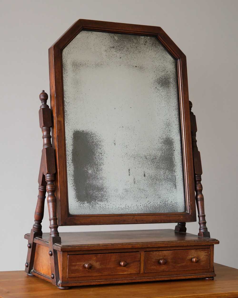 An antique wooden dressing table mirror featuring turned side supports, a two-drawer base with wooden knobs, and an arched mirror with foxed glass.