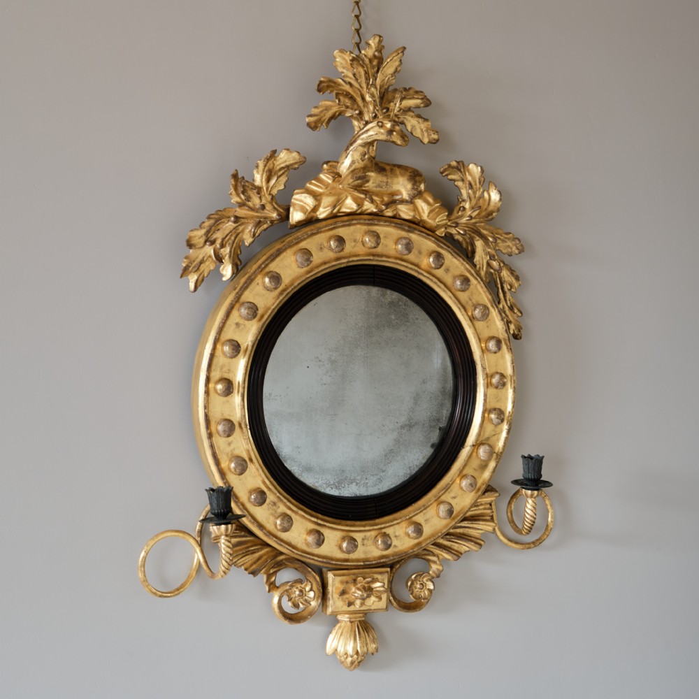 A circular Regency giltwood convex mirror featuring a carved stag cresting, ball ornament frame surround, ebonised reeded slip, and twin candle sconces.