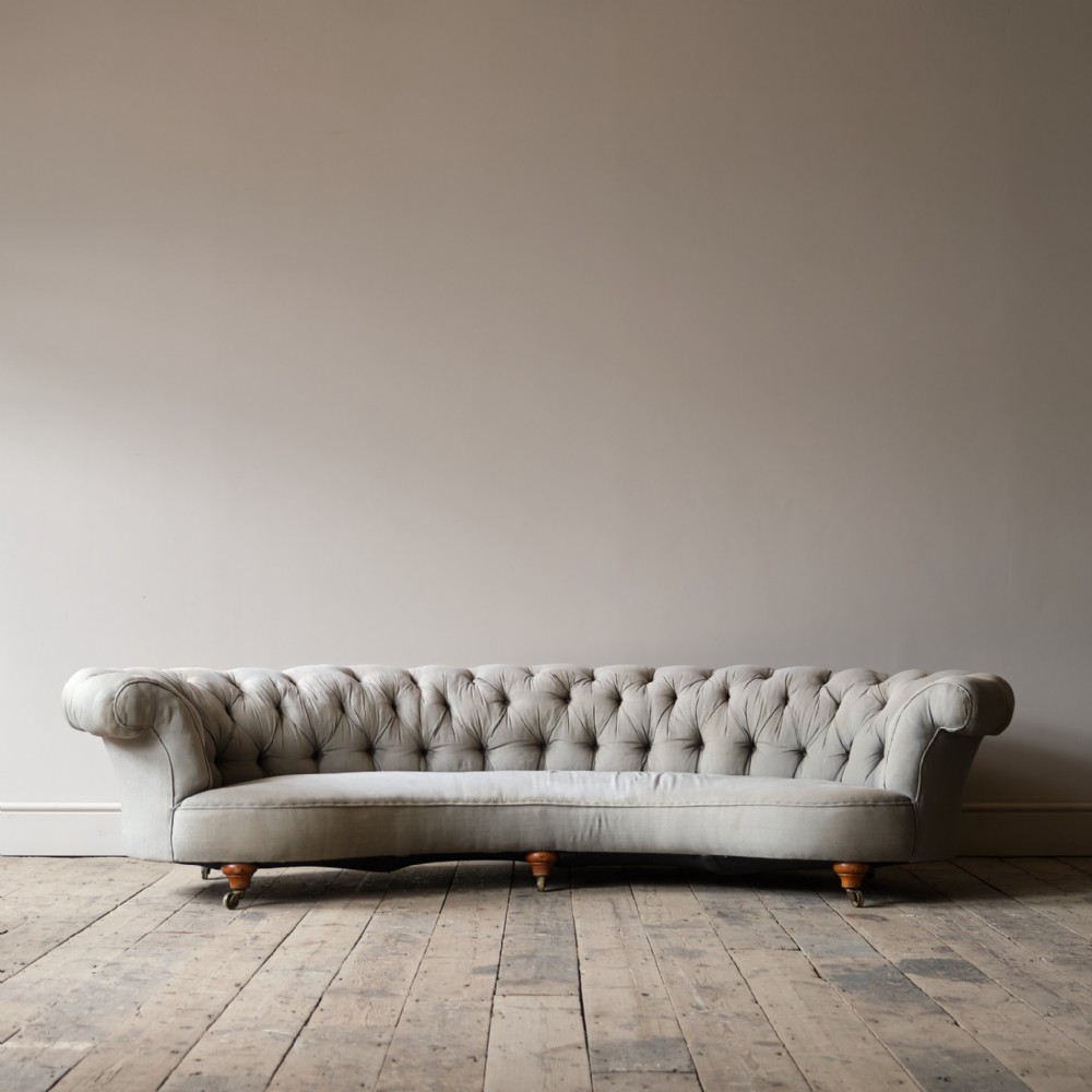 A nineteenth-century Victorian crescent-shaped Chesterfield sofa upholstered in light grey fabric with deep-buttoned back detailing on turned wooden legs with brass castors.