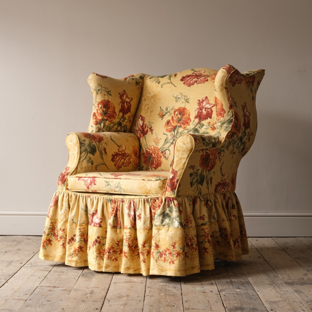 A large George III style wingback armchair upholstered in yellow floral chintz fabric with a pleated skirt.