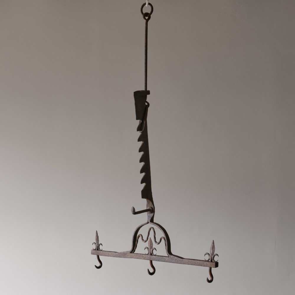 An 18th-century hand-forged wrought iron chimney jack featuring a notched adjustable mechanism, decorative fleur-de-lis motifs, and three hanging hooks.