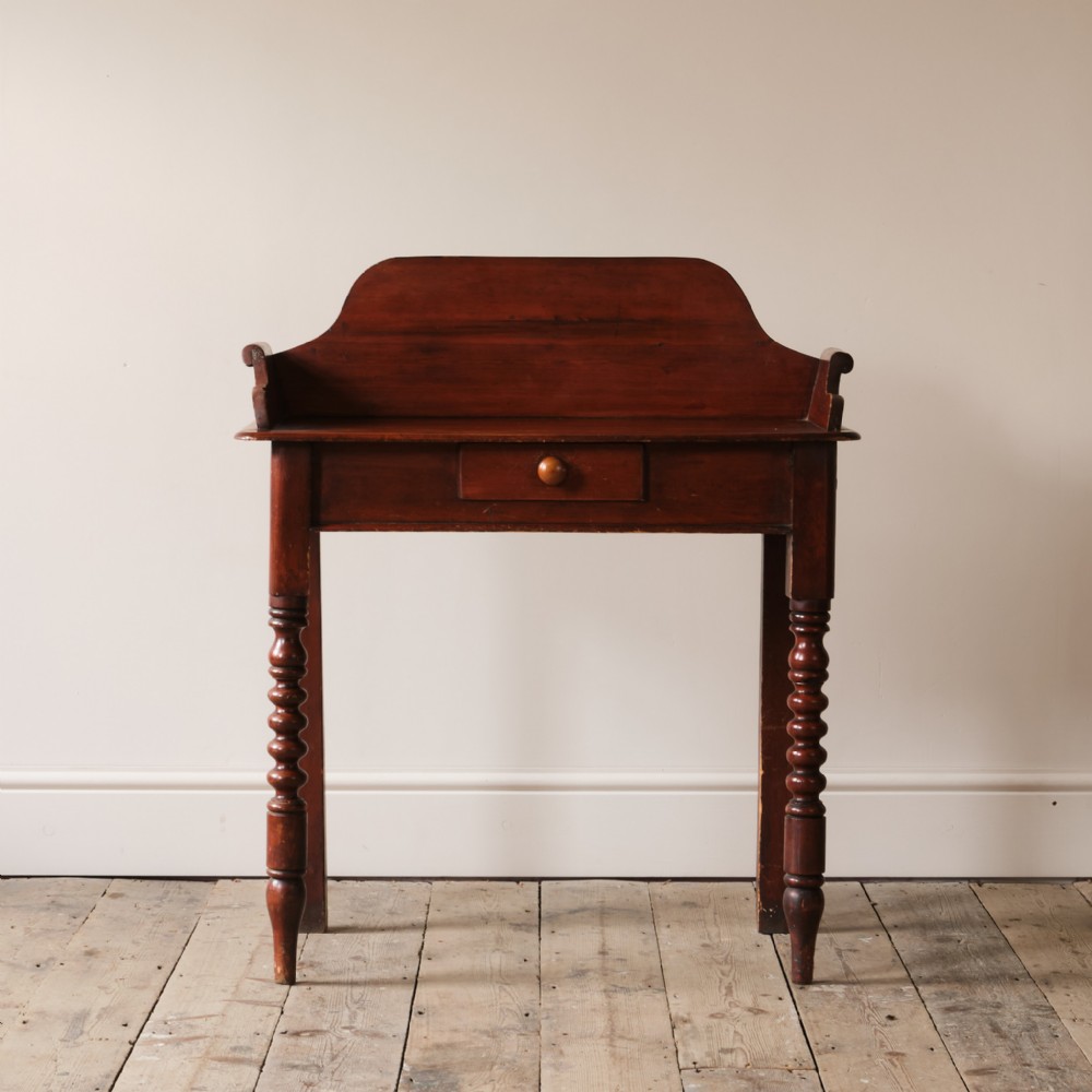 A nineteenth-century pine washstand featuring a raised gallery splashback, a central single drawer with a wooden knob, and turned front legs.