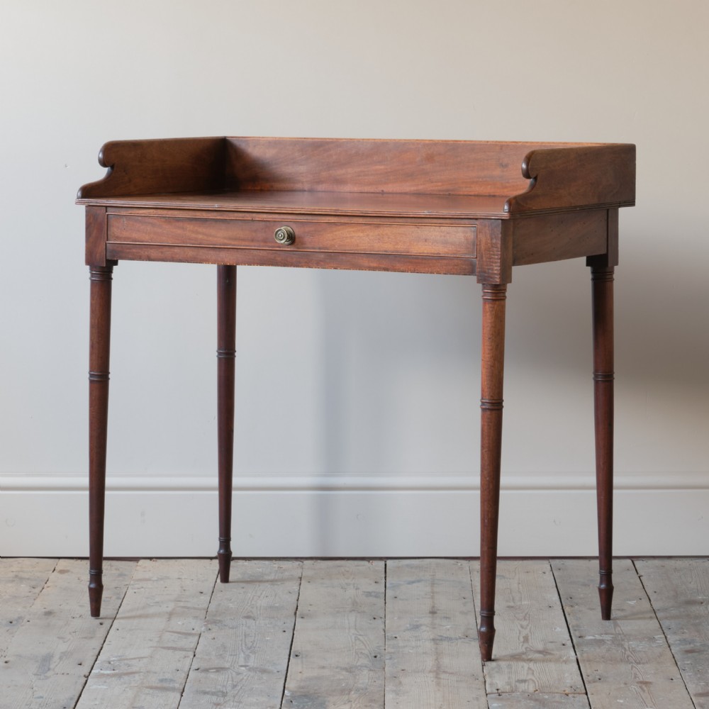 A George III mahogany washstand featuring a low gallery back, single frieze drawer with a brass knob, and four slender turned legs.