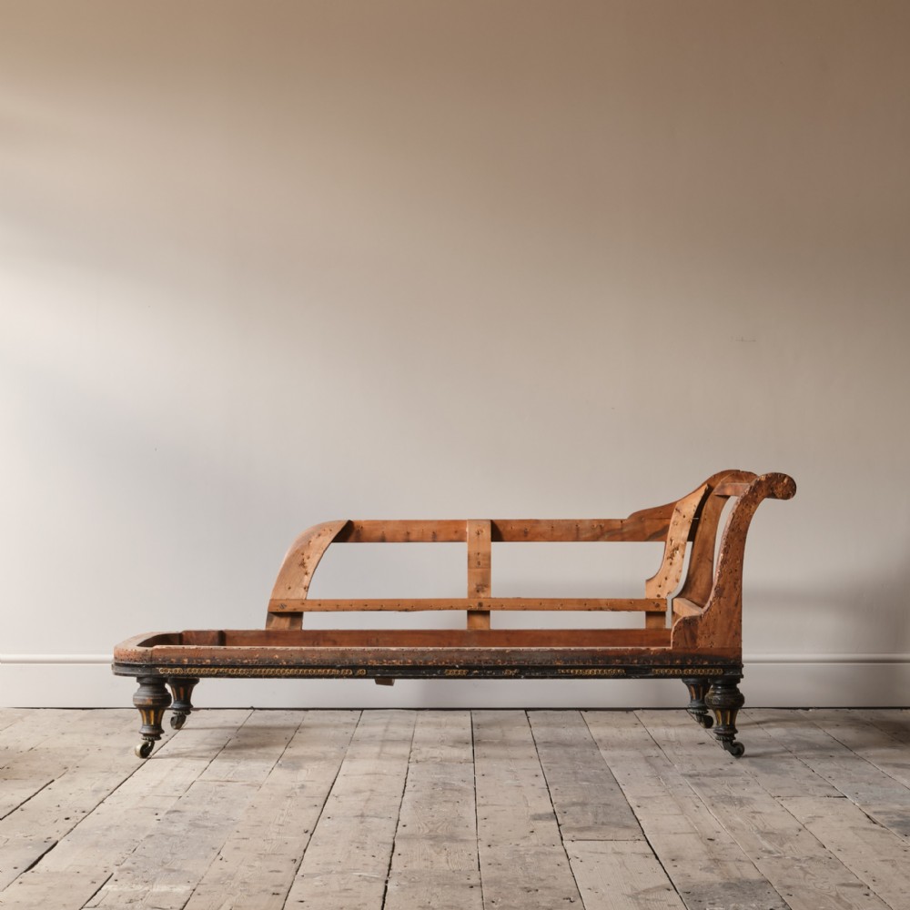 An un-upholstered Victorian wooden chaise longue frame featuring a curved backrest, ebonised seat rail details, and turned legs on brass castors.