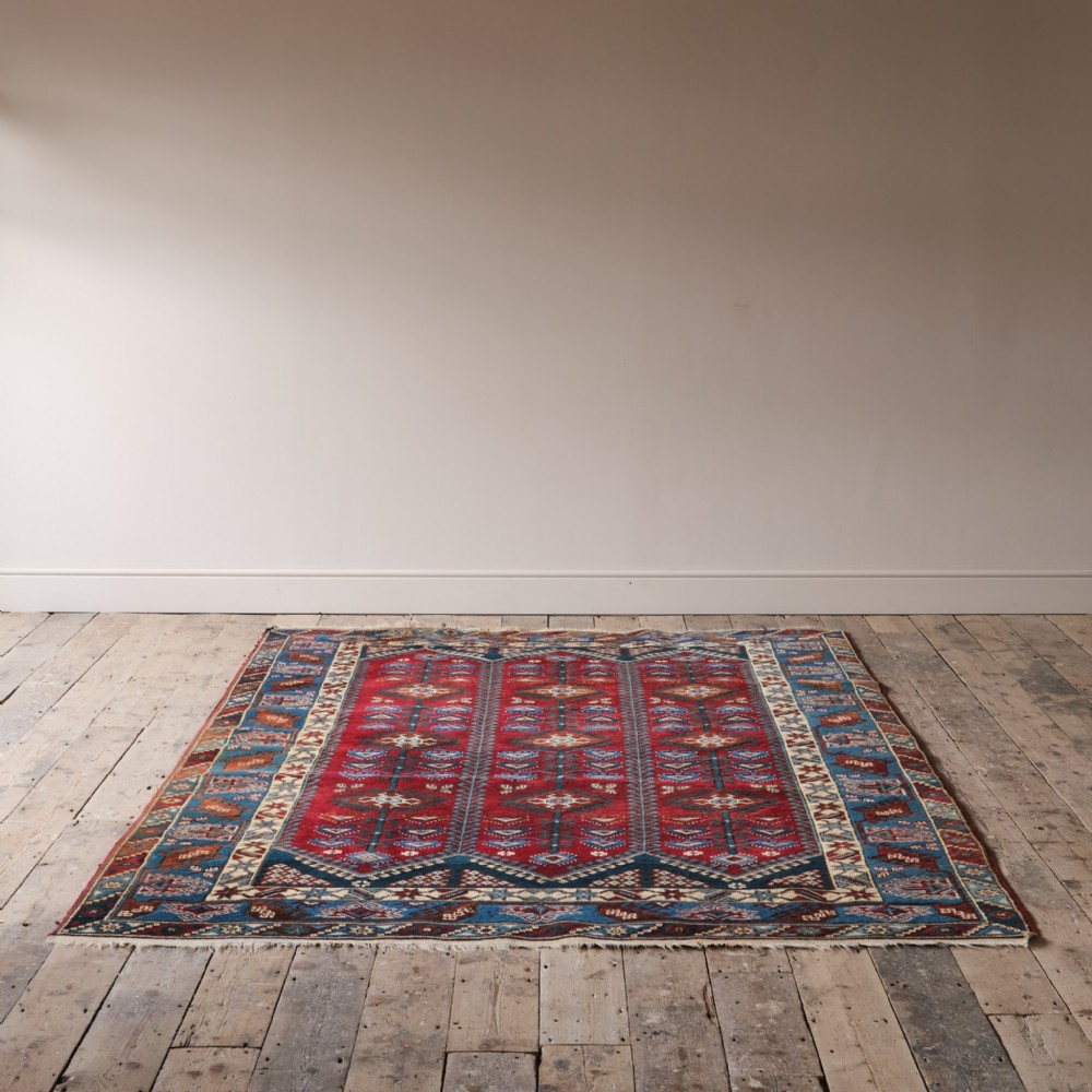 A large hand-knotted wool Shirvan rug featuring a bright red central field with geometric motifs bordered by intricate blue and ivory bands on light timber floorboards.