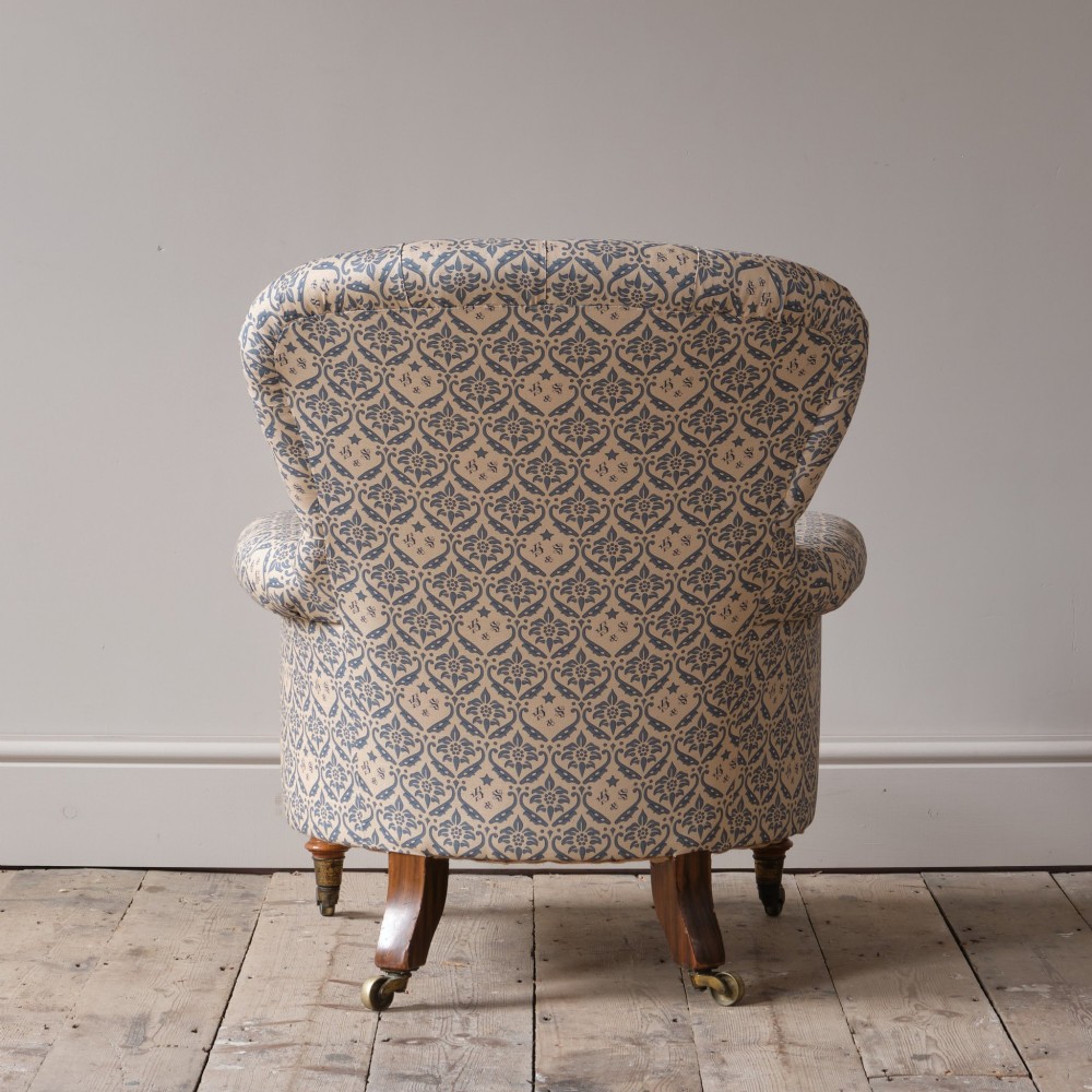 A mid-19th century Victorian upholstered armchair featuring blue and cream patterned fabric, deep button-tufted backrest, turned wooden front legs, and brass castors. View XIII
