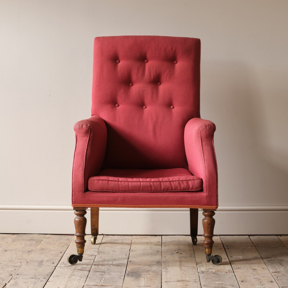 An early 19th-century armchair upholstered in red cloth fabric, featuring a buttoned backrest, padded armrests, and turned timber front legs fitted with brass castors.