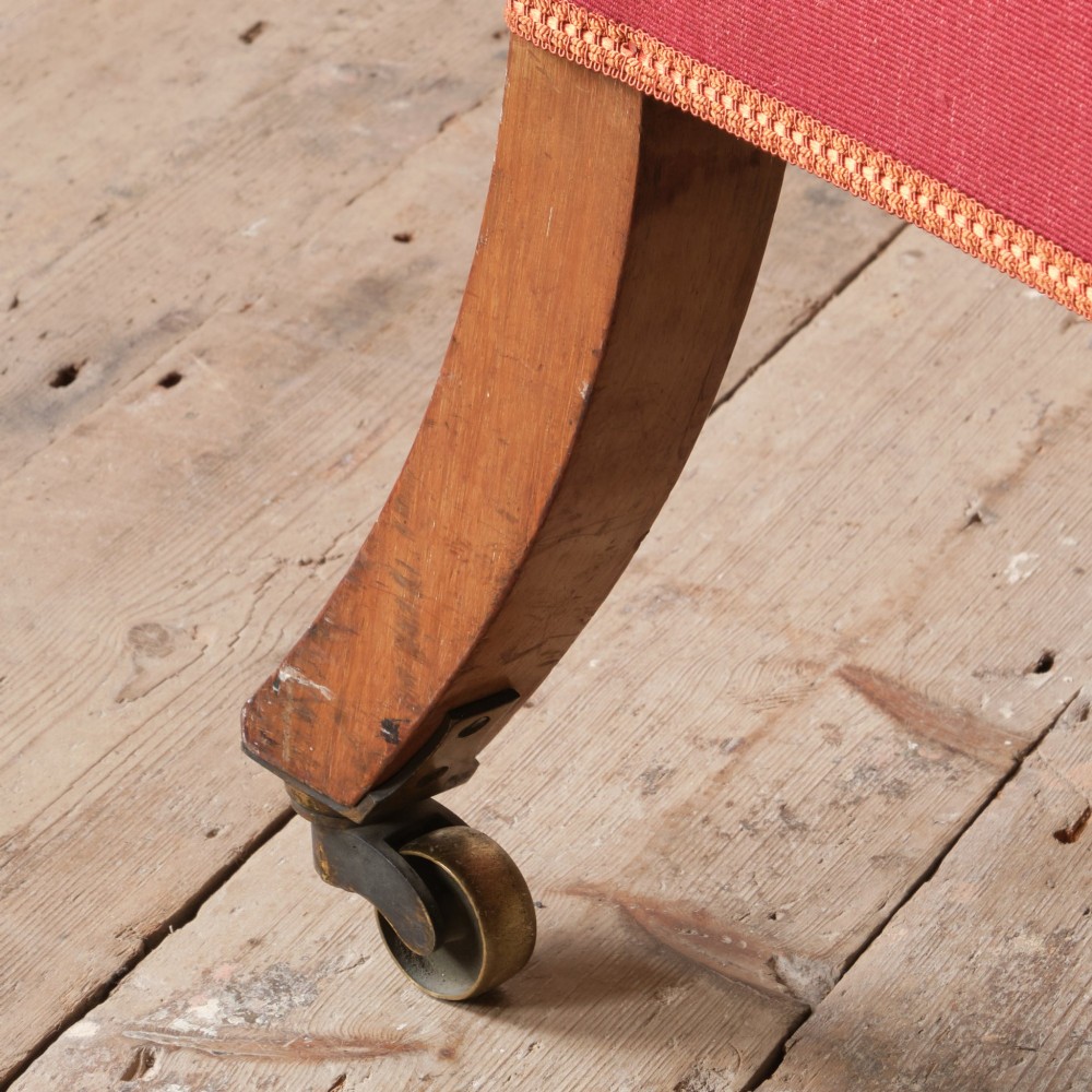 An early 19th-century armchair upholstered in red cloth fabric, featuring a buttoned backrest, padded armrests, and turned timber front legs fitted with brass castors. View VII