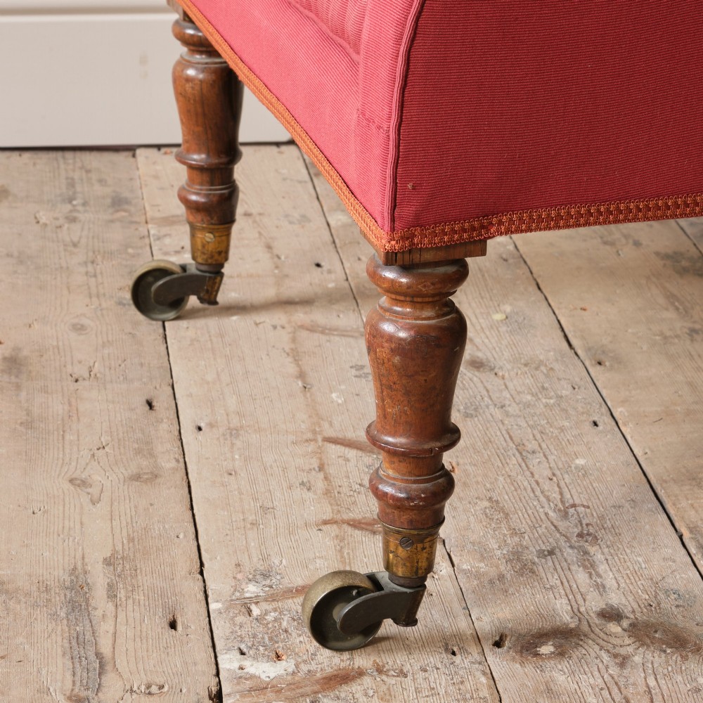An early 19th-century armchair upholstered in red cloth fabric, featuring a buttoned backrest, padded armrests, and turned timber front legs fitted with brass castors. View IX