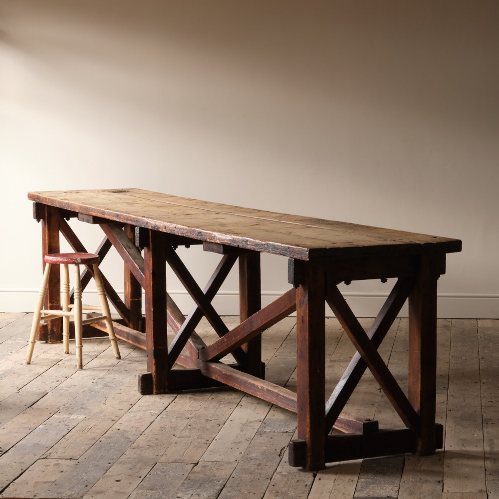 A long antique pine workbench table featuring a heavy plank top supported by a sturdy timber frame with X-frame cross bracing.