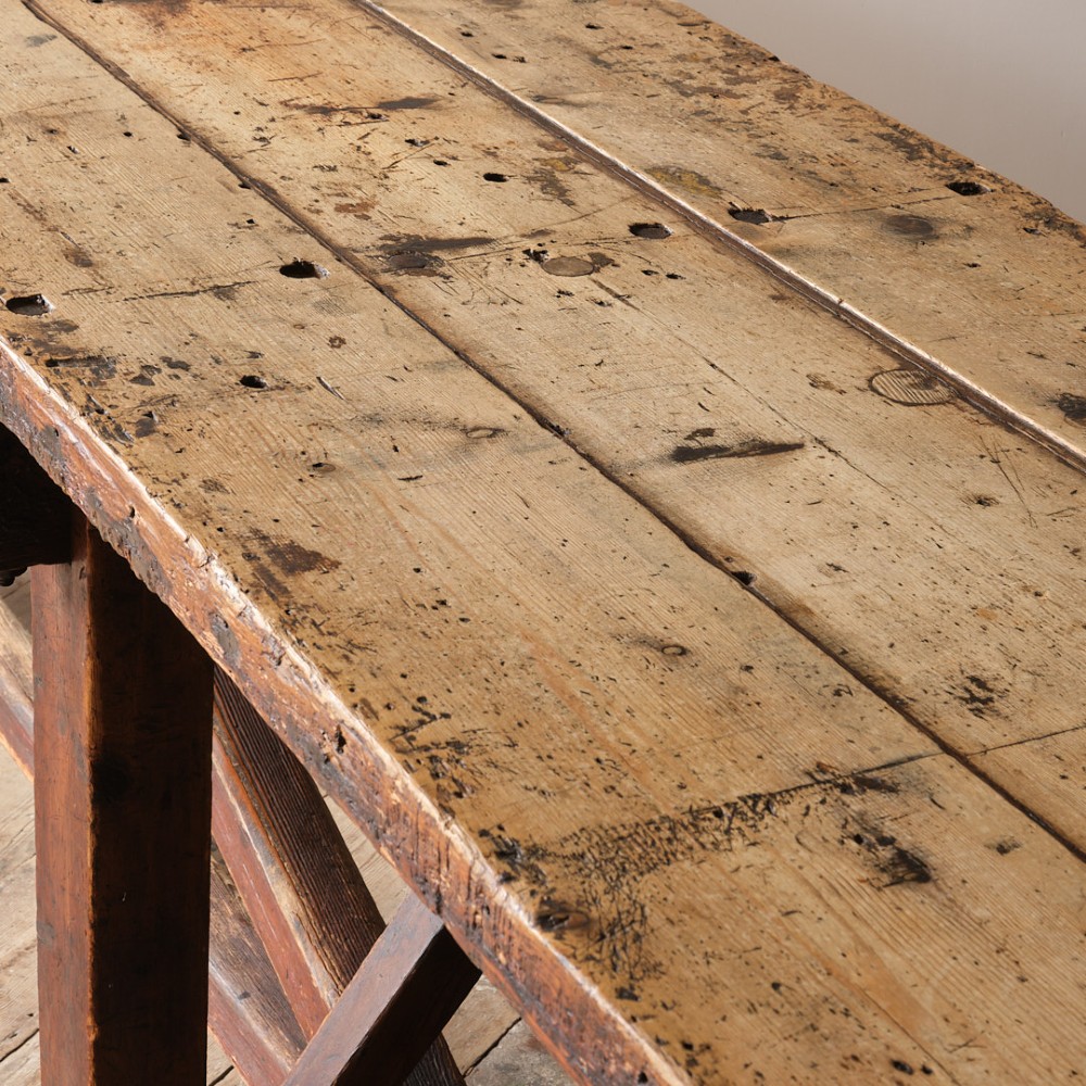 A long antique pine workbench table featuring a heavy plank top supported by a sturdy timber frame with X-frame cross bracing. View X