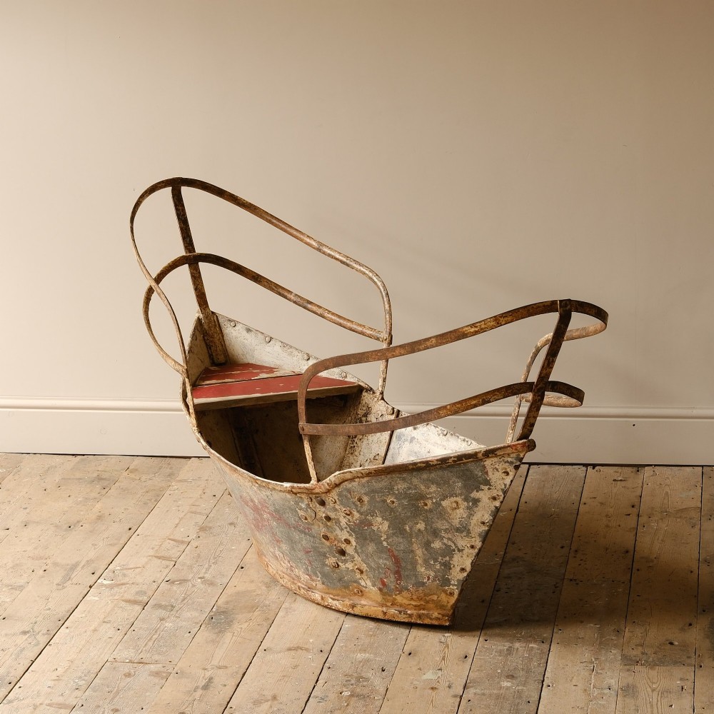 A weathered painted metal fairground swing boat featuring a red wooden seat, riveted sheet body, and curved iron safety rails.