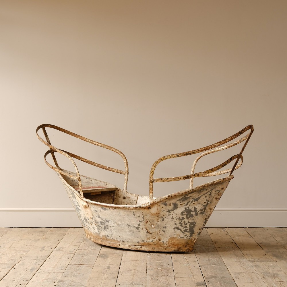 A weathered painted metal fairground swing boat featuring a red wooden seat, riveted sheet body, and curved iron safety rails. View III