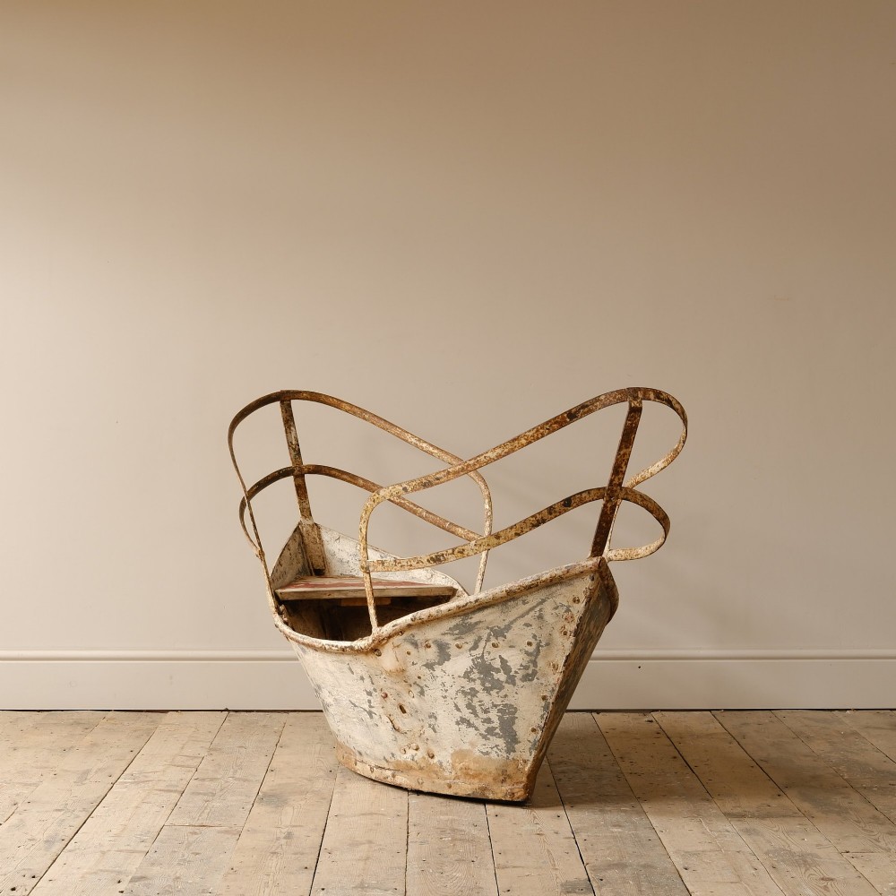 A weathered painted metal fairground swing boat featuring a red wooden seat, riveted sheet body, and curved iron safety rails. View IV