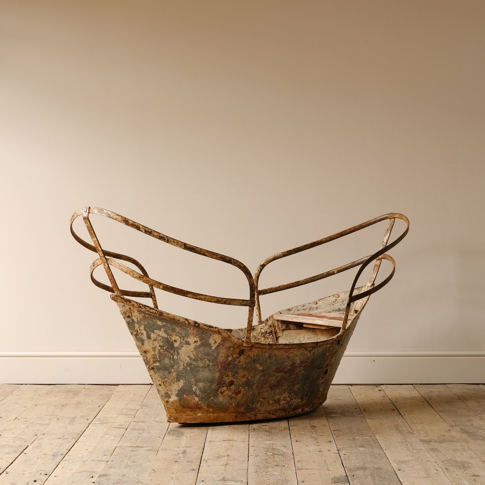 A weathered painted metal fairground swing boat featuring a red wooden seat, riveted sheet body, and curved iron safety rails. View VI