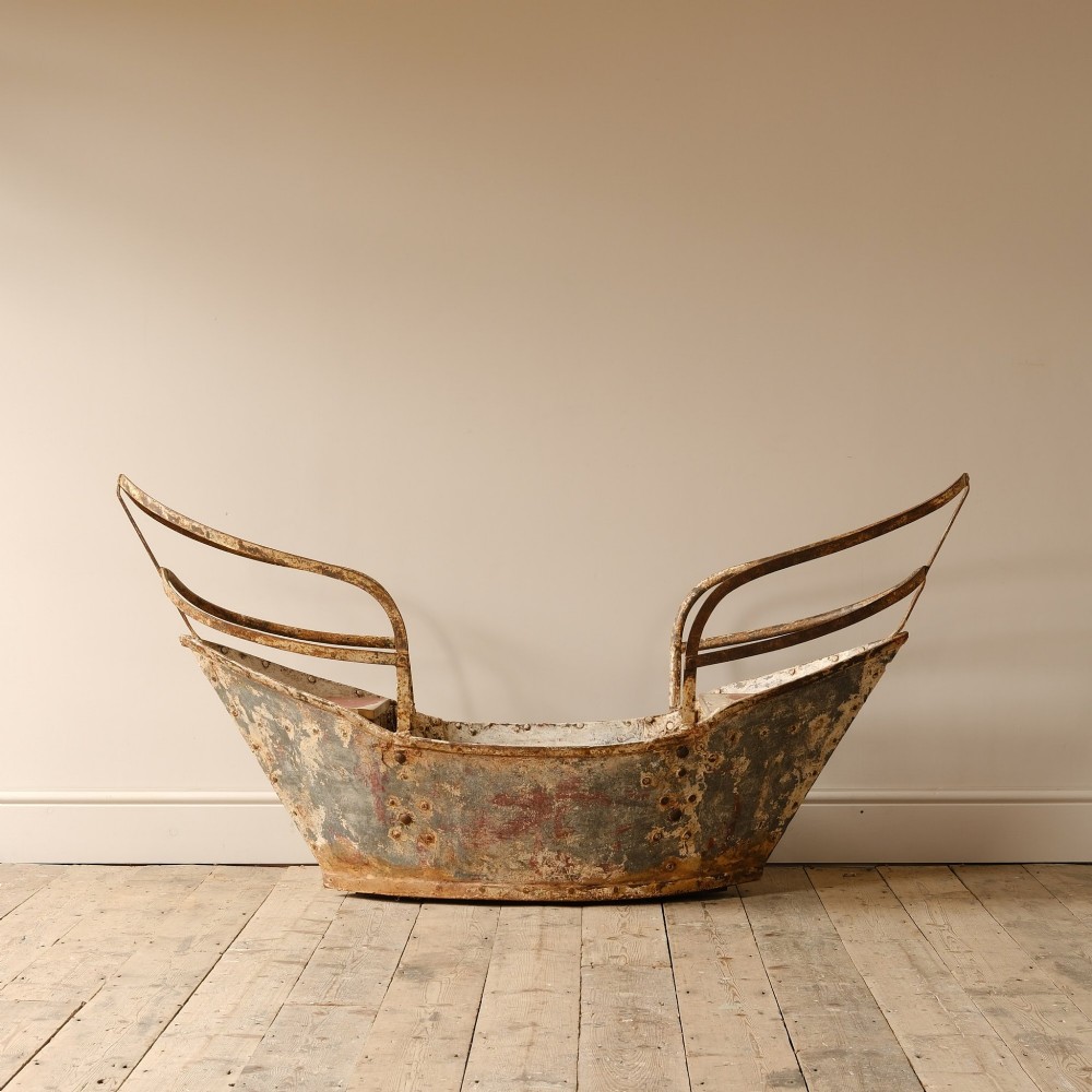 A weathered painted metal fairground swing boat featuring a red wooden seat, riveted sheet body, and curved iron safety rails. View VII