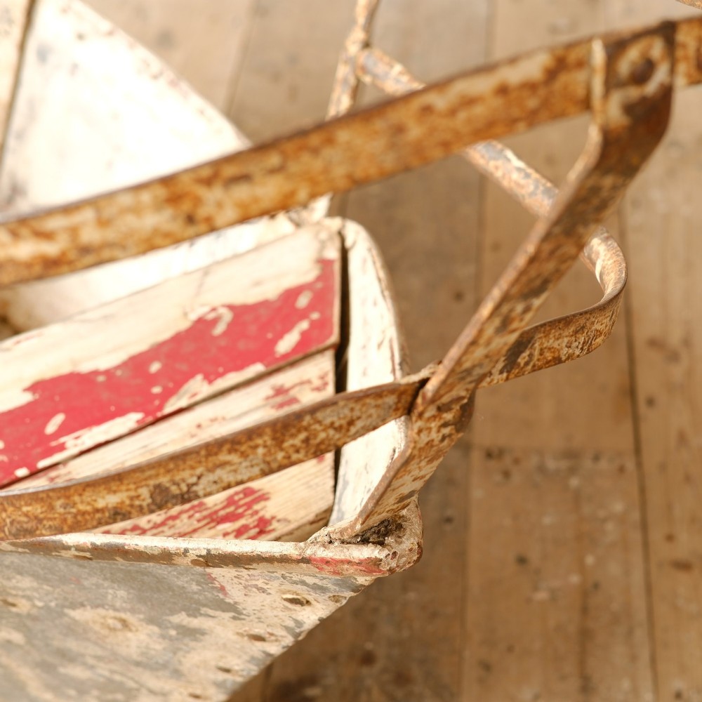 A weathered painted metal fairground swing boat featuring a red wooden seat, riveted sheet body, and curved iron safety rails. View X